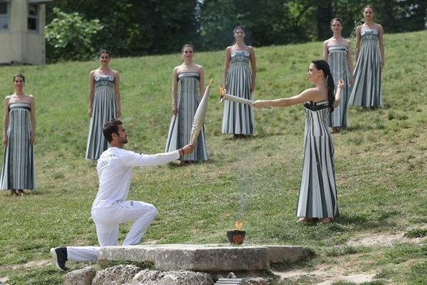 扮演最高女祭司的演員瑪麗·米娜（前右）點(diǎn)燃第一棒火炬手——希臘運(yùn)動(dòng)員、東京奧運(yùn)會(huì)男子賽艇項(xiàng)目金牌得主斯特凡諾斯·恩圖斯科斯手中的火炬。新華社記者 趙丁喆 攝